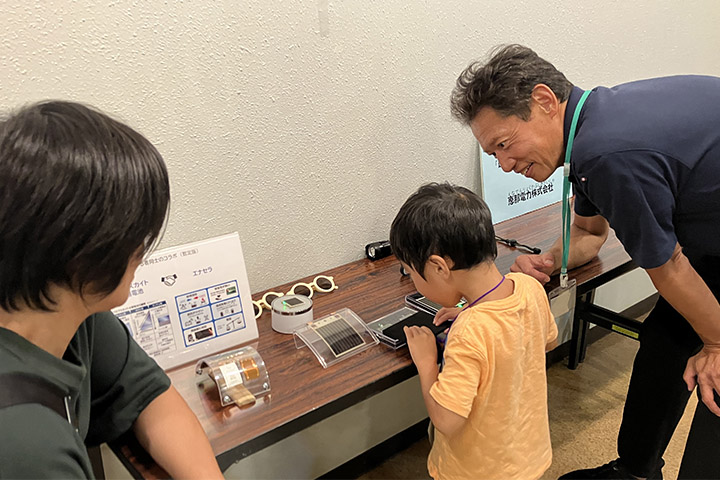 ブースを訪れた子にペロブスカイト太陽電池の説明をするエネコートテクノロジーズ社の社員(右)。薄くて曲げられる電池に興味津々の様子